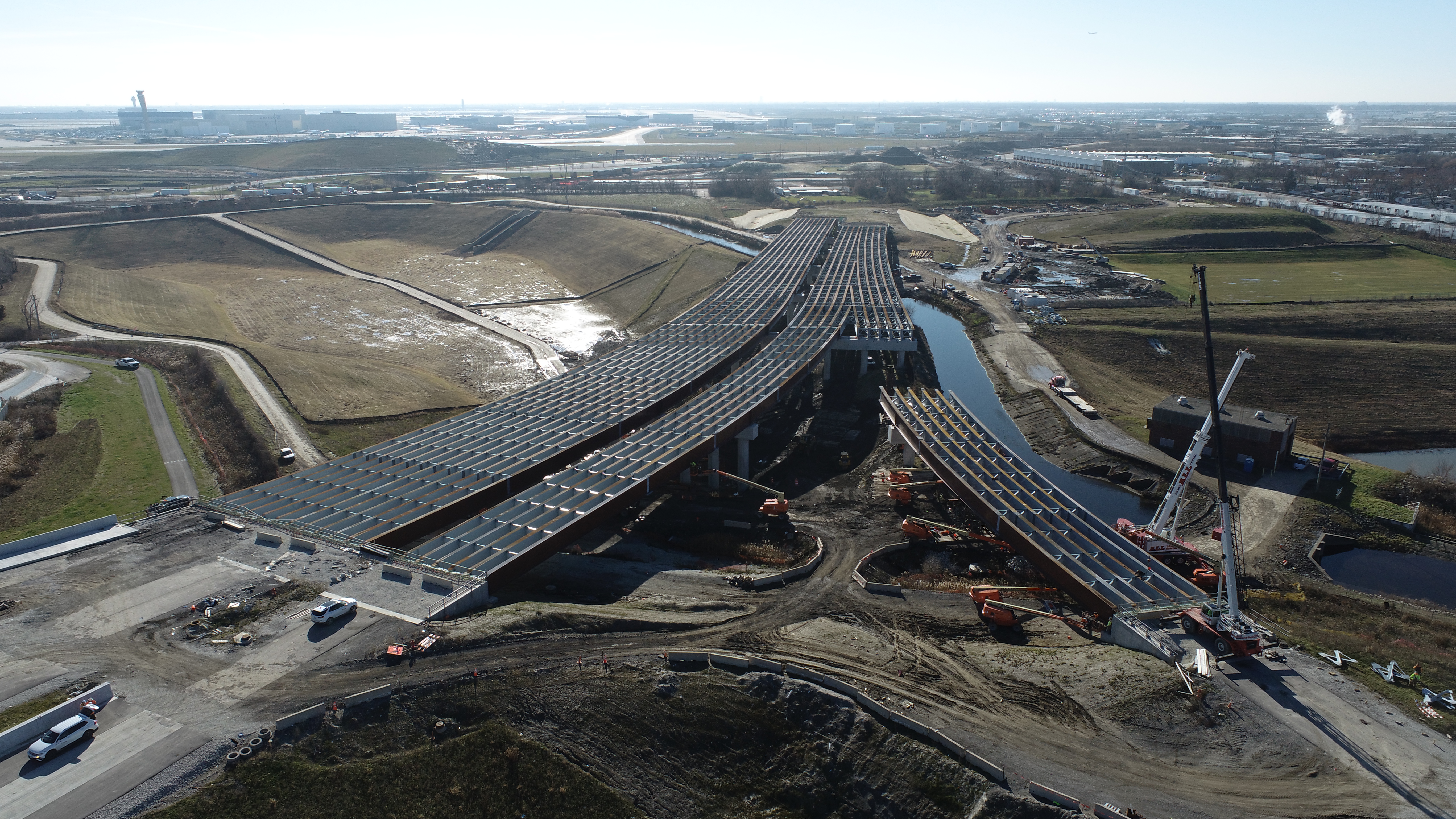 Aerial view of a highway bridge under construction over a canal, with construction equipment and cranes visible, surrounded by open land near an urban area